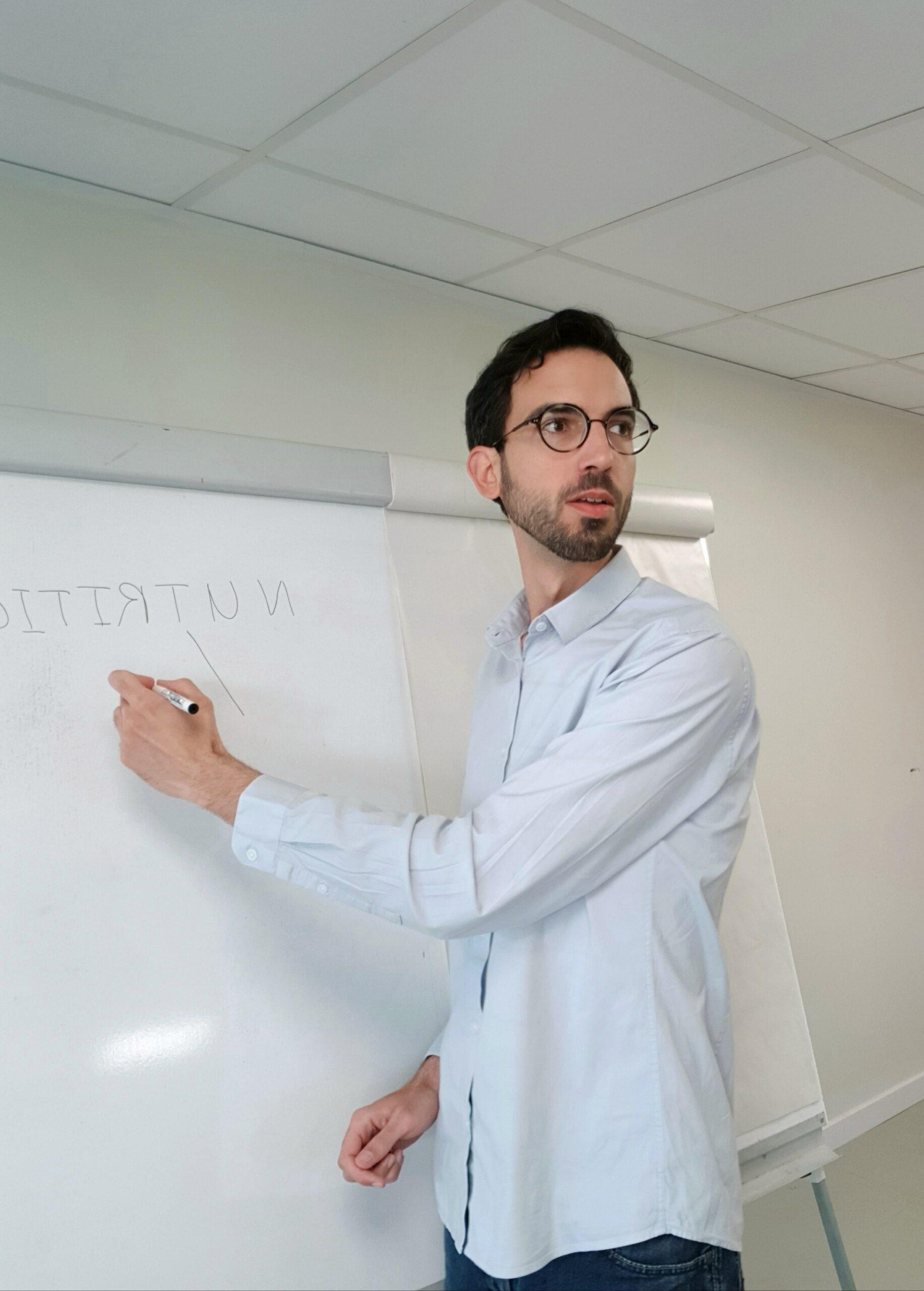 Un homme avec une chemise grise et des lunettes rondes donne un cours devant un tableau blanc.
